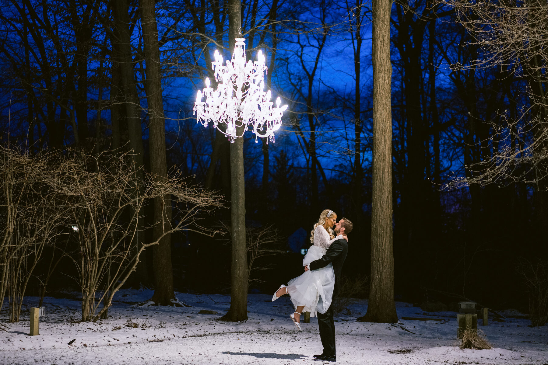 Groom lifting bride beneath a chandelier in a twilight forest